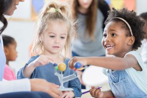 Diverse female preschool students touch a solar system model. Their teacher is teaching them about the planets.