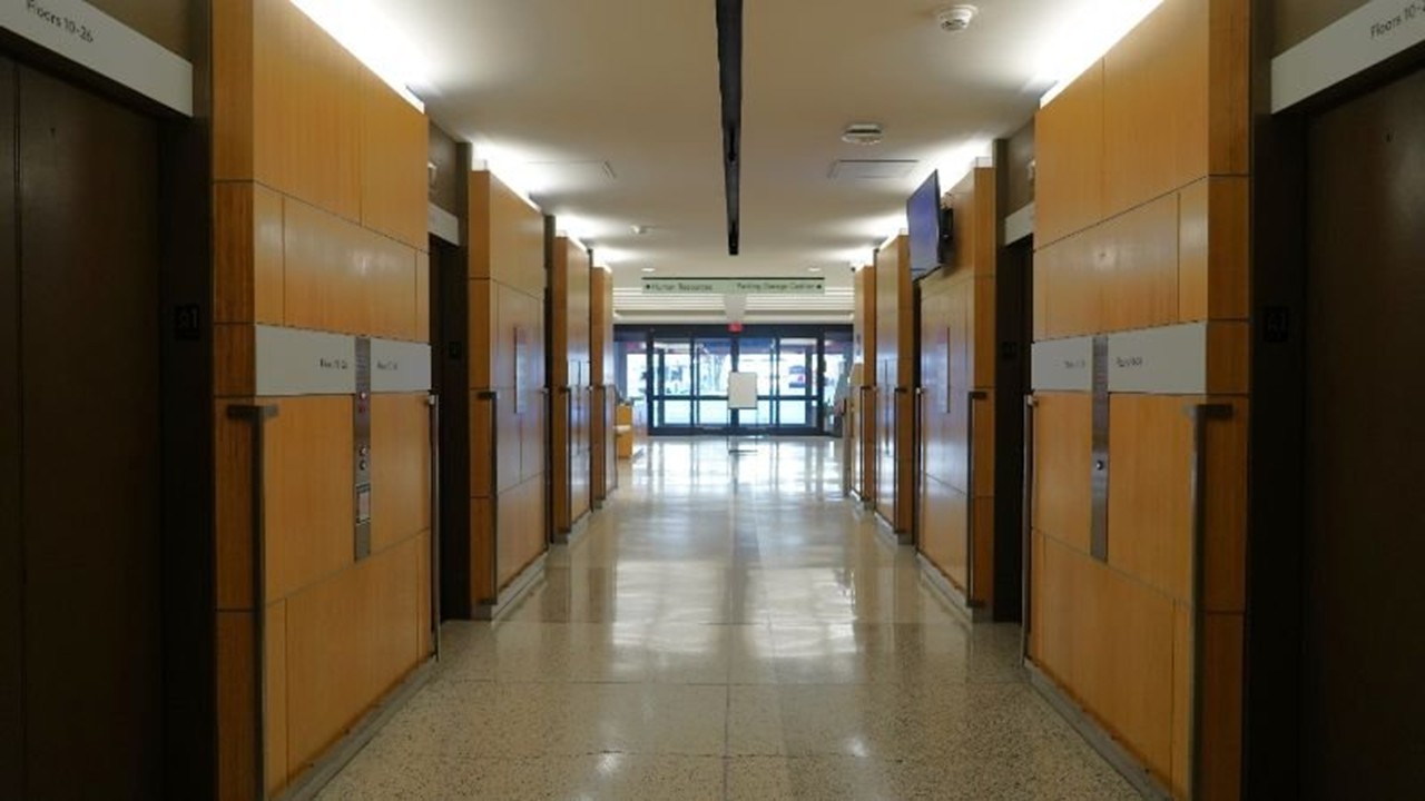 A wide hallway lined with wooden elevator doors on both sides leads toward glass exit doors in the distance. Overhead lighting reflects on the polished floor.