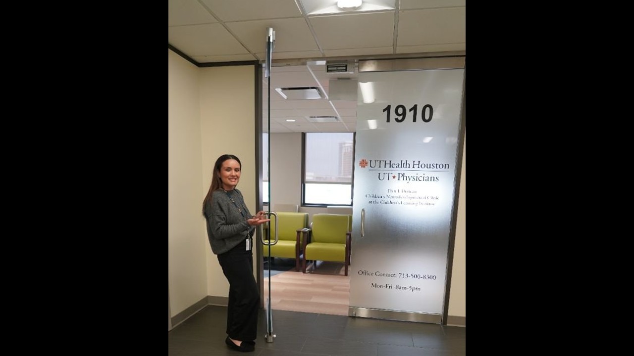 A person stands beside a glass door labeled “1910” and “UTHealth Houston UT Physicians,” holding the door open to reveal a waiting area with green chairs. The space is brightly lit with large windows in the background.