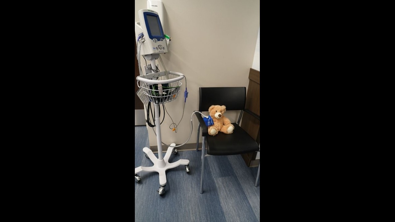 A blood‑pressure monitor on a wheeled stand is positioned next to a black chair, where a stuffed teddy bear sits with a pediatric blood‑pressure cuff wrapped around its arm. The setting appears to be an exam room with blue flooring and a plain wall.
