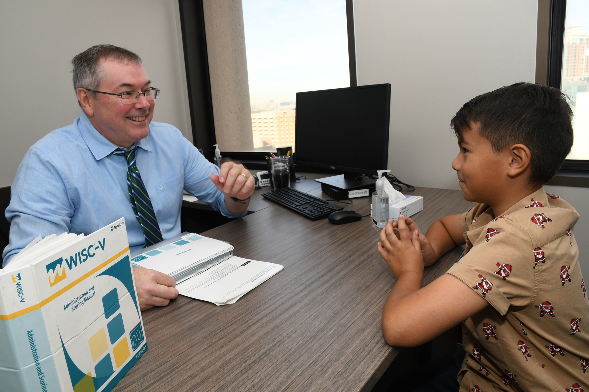Adult clinician administers a cognitive assessment to a child at a desk, with testing materials visible.