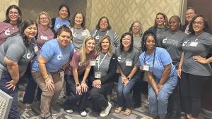 “A large group of staff members poses for a photo in a conference room with patterned walls. Some individuals stand in the back row while others kneel or sit in front. Many participants wear matching or similar event T-shirts and name badges. The group faces the camera in a close formation, suggesting a team or cohort attending a shared professional learning event.”